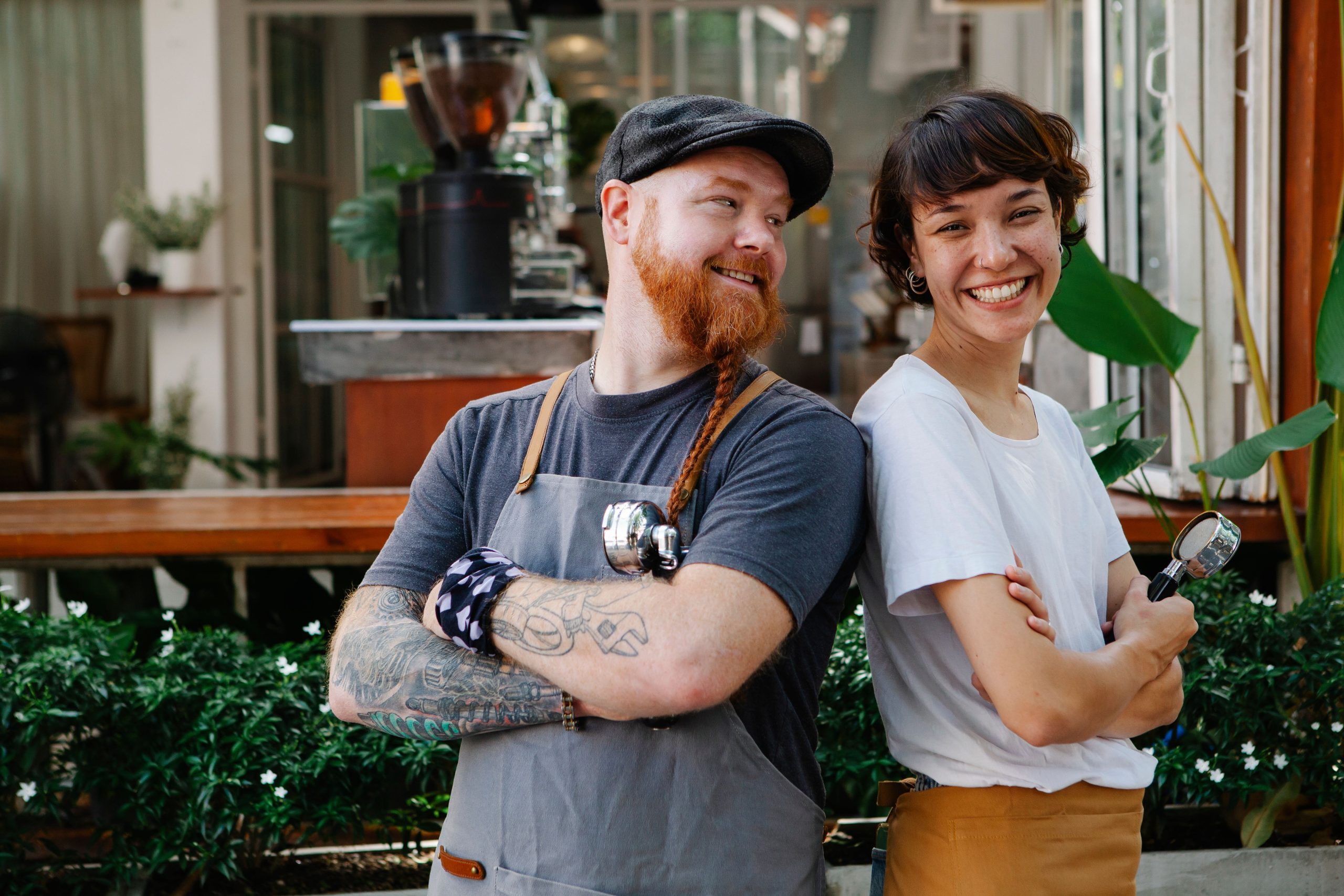 Two baristas – a male and a female standing shoulder to shoulder in front of a coffee shop. They are smiling.  